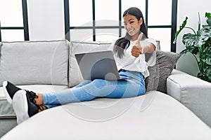 Young hispanic woman using laptop at home sitting on the sofa smiling happy and positive, thumb up doing excellent and approval