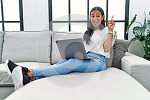 Young hispanic woman using laptop at home sitting on the sofa surprised with an idea or question pointing finger with happy face,