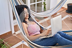 Young hispanic woman using laptop lying on hammock at home terrace