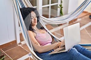 Young hispanic woman using laptop lying on hammock at home terrace
