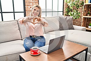 Young hispanic woman using laptop sitting on the sofa at home smiling in love doing heart symbol shape with hands