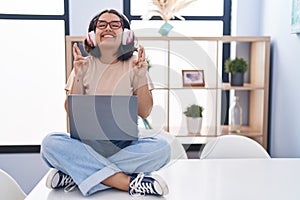 Young hispanic woman using laptop sitting on the table wearing headphones gesturing finger crossed smiling with hope and eyes