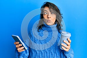Young hispanic woman using smartphone and drinking a cup of coffee making fish face with mouth and squinting eyes, crazy and
