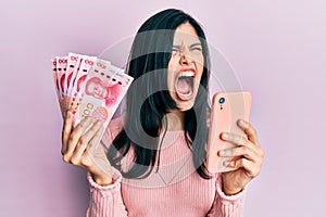 Young hispanic woman using smartphone holding chinese yuan banknotes angry and mad screaming frustrated and furious, shouting with