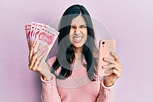 Young hispanic woman using smartphone holding chinese yuan banknotes clueless and confused expression