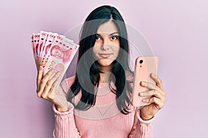 Young hispanic woman using smartphone holding chinese yuan banknotes relaxed with serious expression on face