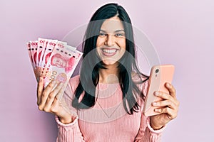 Young hispanic woman using smartphone holding chinese yuan banknotes smiling with a happy and cool smile on face