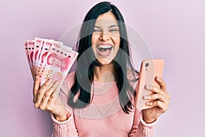 Young hispanic woman using smartphone holding chinese yuan banknotes smiling and laughing hard out loud because funny crazy joke