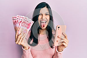 Young hispanic woman using smartphone holding chinese yuan banknotes sticking tongue out happy with funny expression