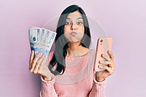 Young hispanic woman using smartphone holding colombian pesos banknotes puffing cheeks with funny face
