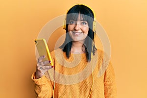 Young hispanic woman using smartphone wearing headphones looking positive and happy standing and smiling with a confident smile