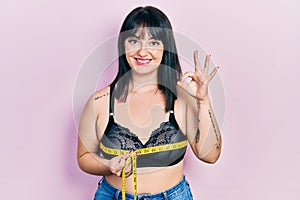 Young hispanic woman using tape measure measuring breast doing ok sign with fingers, smiling friendly gesturing excellent symbol