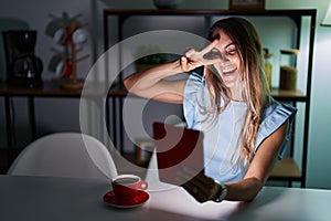 Young hispanic woman using touchpad sitting on the table at night doing peace symbol with fingers over face, smiling cheerful
