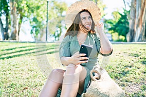 Young hispanic woman on vacation smiling happy sitting on the grass using smartphone at park