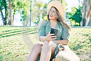 Young hispanic woman on vacation smiling happy sitting on the grass using smartphone at park
