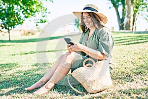 Young hispanic woman on vacation smiling happy sitting on the grass using smartphone at park