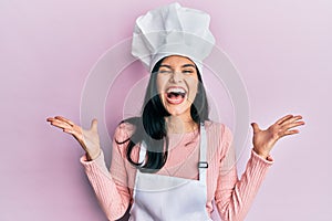 Young hispanic woman wearing baker uniform and cook hat celebrating mad and crazy for success with arms raised and closed eyes