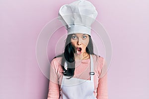 Young hispanic woman wearing baker uniform and cook hat scared and amazed with open mouth for surprise, disbelief face