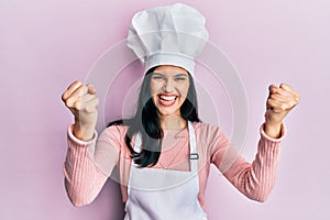 Young hispanic woman wearing baker uniform and cook hat screaming proud, celebrating victory and success very excited with raised