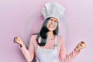 Young hispanic woman wearing baker uniform and cook hat very happy and excited doing winner gesture with arms raised, smiling and