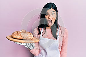Young hispanic woman wearing baker uniform holding homemade bread scared and amazed with open mouth for surprise, disbelief face