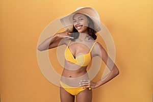 Young hispanic woman wearing bikini and summer hat smiling doing phone gesture with hand and fingers like talking on the telephone