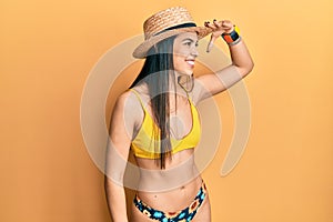 Young hispanic woman wearing bikini and summer hat very happy and smiling looking far away with hand over head