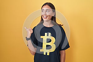 Young hispanic woman wearing bitcoin t shirt smiling with happy face looking and pointing to the side with thumb up