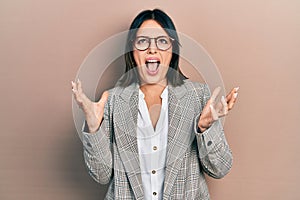 Young hispanic woman wearing business clothes and glasses crazy and mad shouting and yelling with aggressive expression and arms
