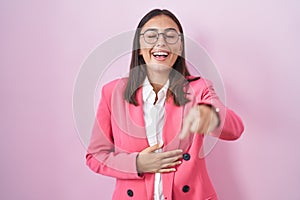 Young hispanic woman wearing business clothes and glasses laughing at you, pointing finger to the camera with hand over body,