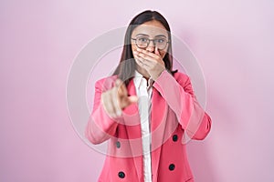 Young hispanic woman wearing business clothes and glasses laughing at you, pointing finger to the camera with hand over mouth,