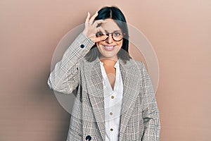 Young hispanic woman wearing business clothes and glasses smiling happy doing ok sign with hand on eye looking through fingers