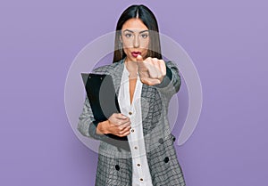 Young hispanic woman wearing business clothes holding clipboard pointing with finger to the camera and to you, confident gesture