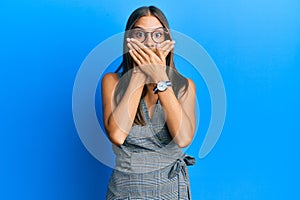Young hispanic woman wearing business dress and glasses shocked covering mouth with hands for mistake