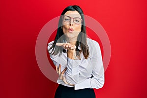 Young hispanic woman wearing business shirt and glasses looking at the camera blowing a kiss with hand on air being lovely and