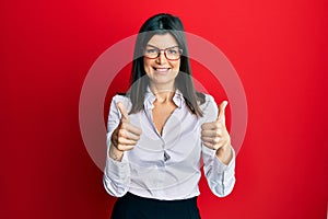 Young hispanic woman wearing business shirt and glasses success sign doing positive gesture with hand, thumbs up smiling and happy