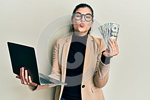 Young hispanic woman wearing business style holding laptop and 50 euros looking at the camera blowing a kiss being lovely and sexy