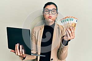 Young hispanic woman wearing business style holding laptop and 50 euros puffing cheeks with funny face