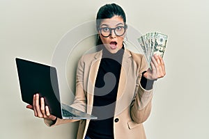Young hispanic woman wearing business style holding laptop and 50 euros in shock face, looking skeptical and sarcastic, surprised