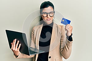 Young hispanic woman wearing business style holding laptop and credit card clueless and confused expression