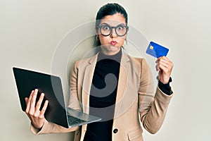 Young hispanic woman wearing business style holding laptop and credit card puffing cheeks with funny face