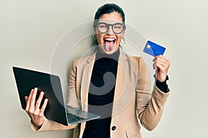 Young hispanic woman wearing business style holding laptop and credit card sticking tongue out happy with funny expression