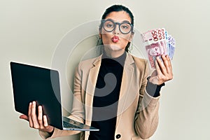 Young hispanic woman wearing business style holding laptop and swedish krone looking at the camera blowing a kiss being lovely and