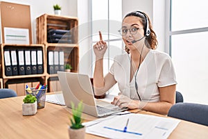 Young hispanic woman wearing call center agent headset at the office pointing finger up with successful idea