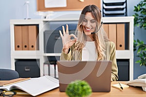 Young hispanic woman wearing call center agent headset working at the office doing ok sign with fingers, smiling friendly