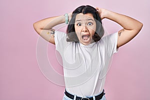 Young hispanic woman wearing casual white t shirt over pink background crazy and scared with hands on head, afraid and surprised