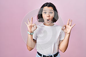 Young hispanic woman wearing casual white t shirt over pink background looking surprised and shocked doing ok approval symbol with