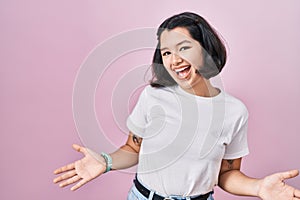 Young hispanic woman wearing casual white t shirt over pink background smiling cheerful with open arms as friendly welcome,