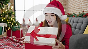 Young hispanic woman wearing christmas hat unpacking gift with surprised face at home