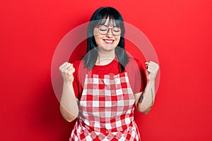 Young hispanic woman wearing cook apron and glasses excited for success with arms raised and eyes closed celebrating victory